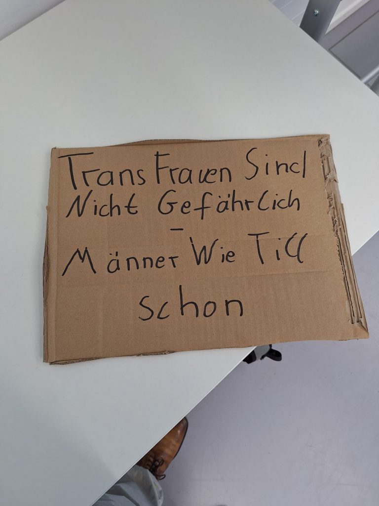 Pappschild, auf dem Tisch liegend. Darauf steht: "Transfrauen sind nicht gefährlich - Männer wie Till schon."